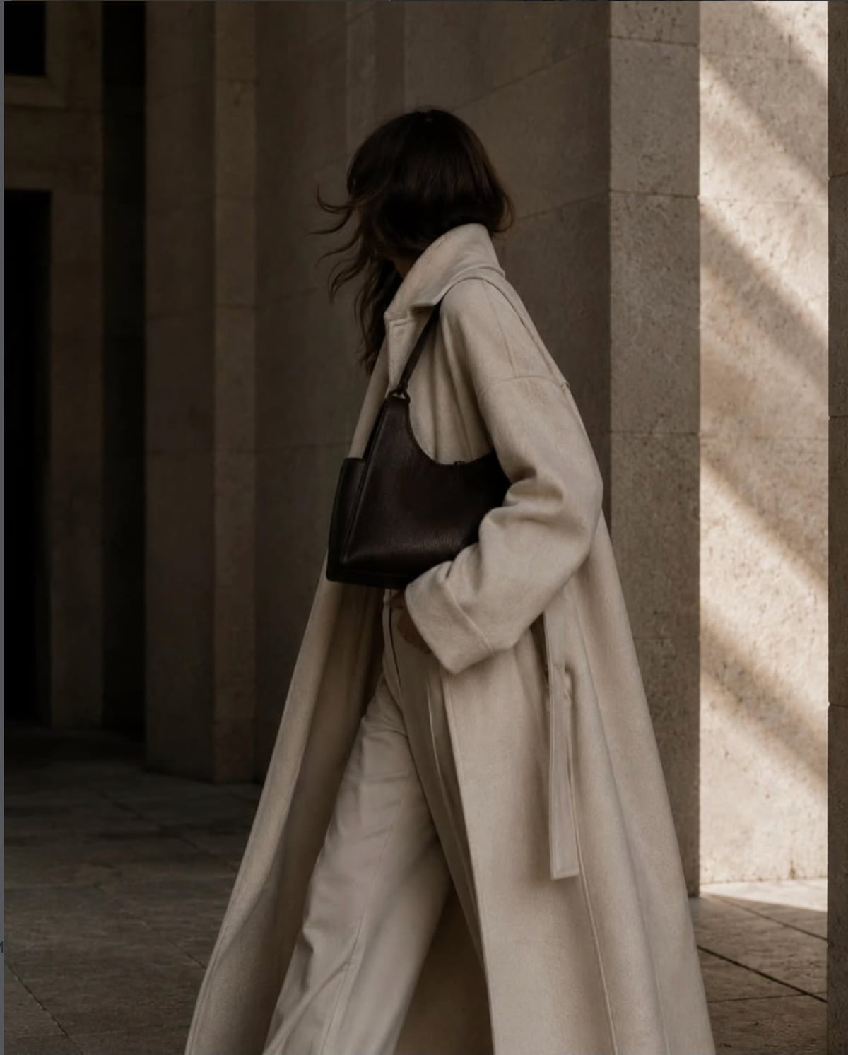 Editorial street portrait — a woman in a long cream wool coat carries a dark leather shoulder bag past stone columns in raking light.