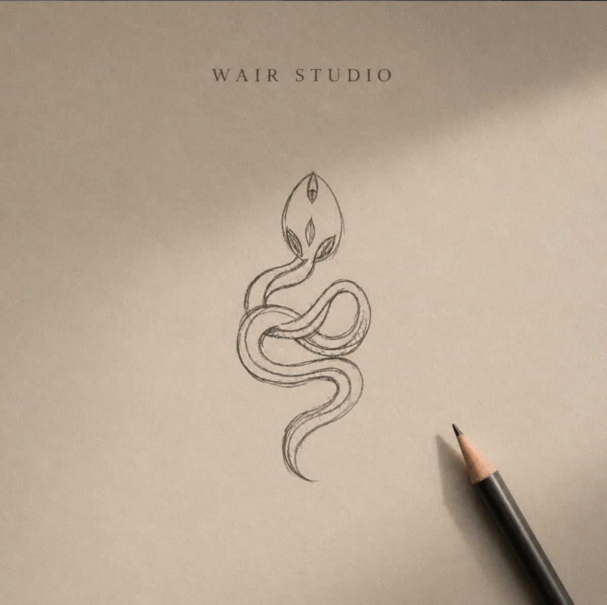 Pencil sketch of a coiled serpent emblem on cream paper, the pencil resting beside the drawing on the WAIR Studio workbench.
