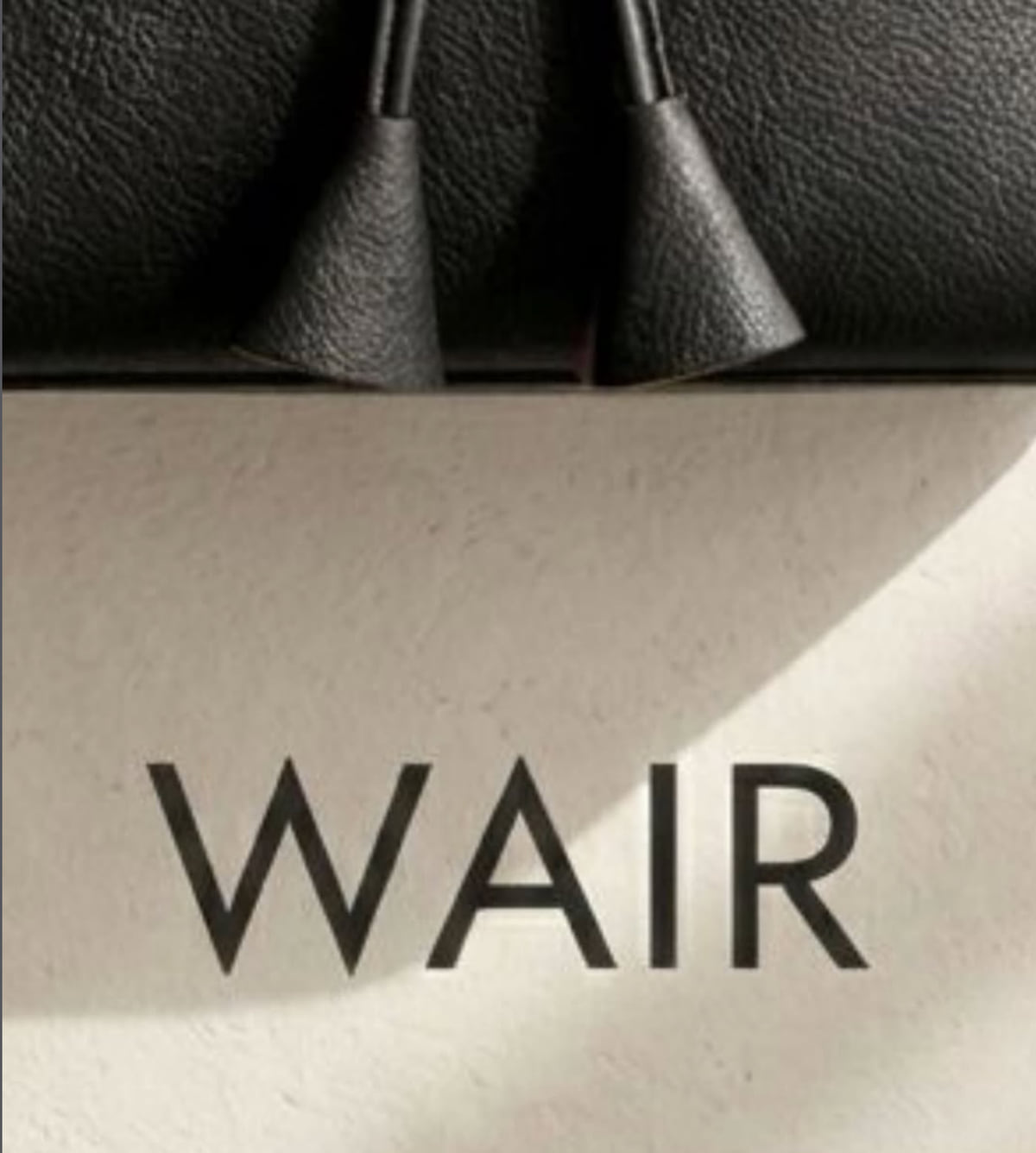 Close-up of two leather handles on a dark bag with the WAIR wordmark in a serif typeface on a cream wall below.