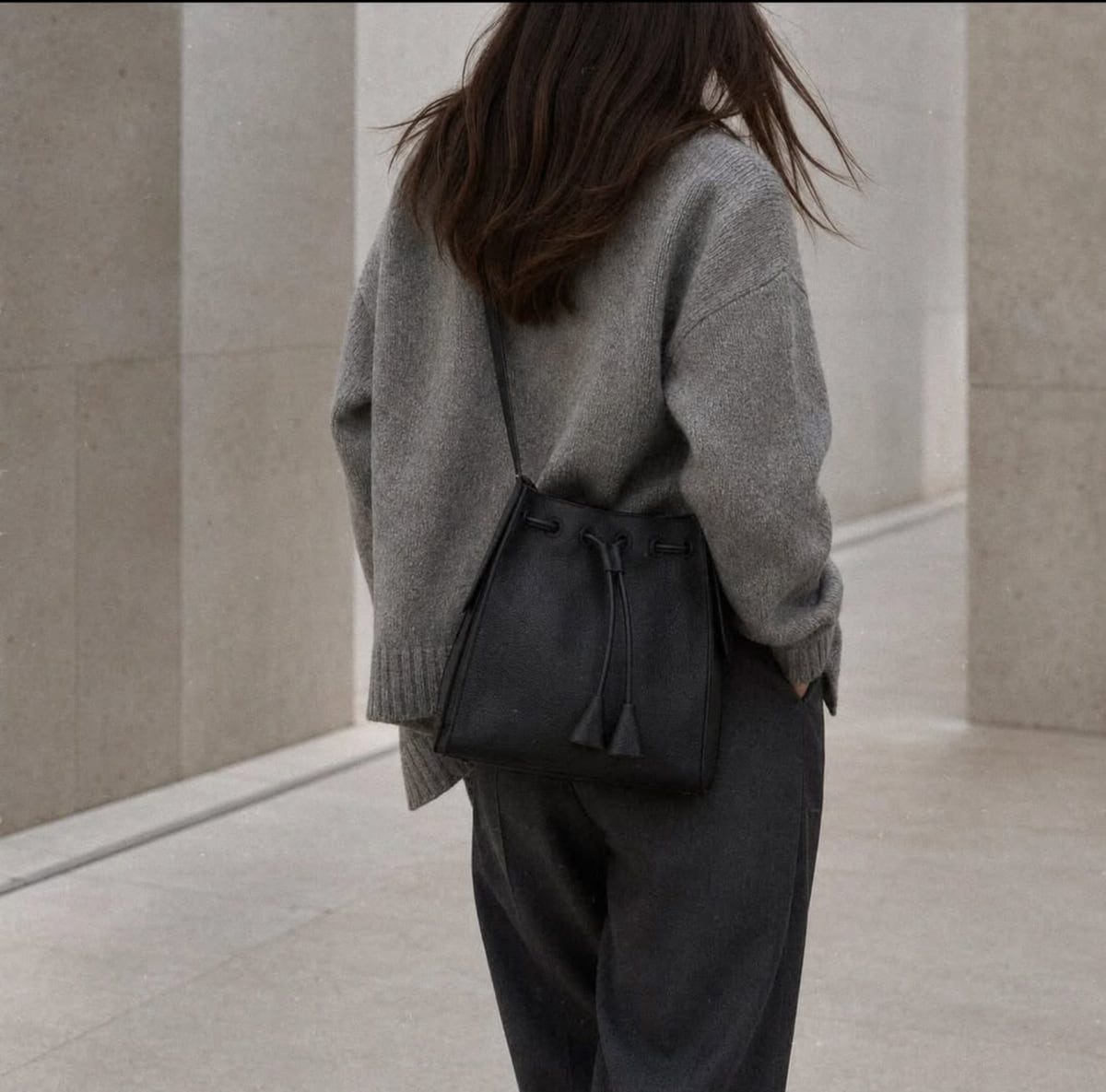 A woman in a gray wool sweater and dark trousers, seen from behind, carries a black drawstring bucket bag on her shoulder against a pale stone wall.