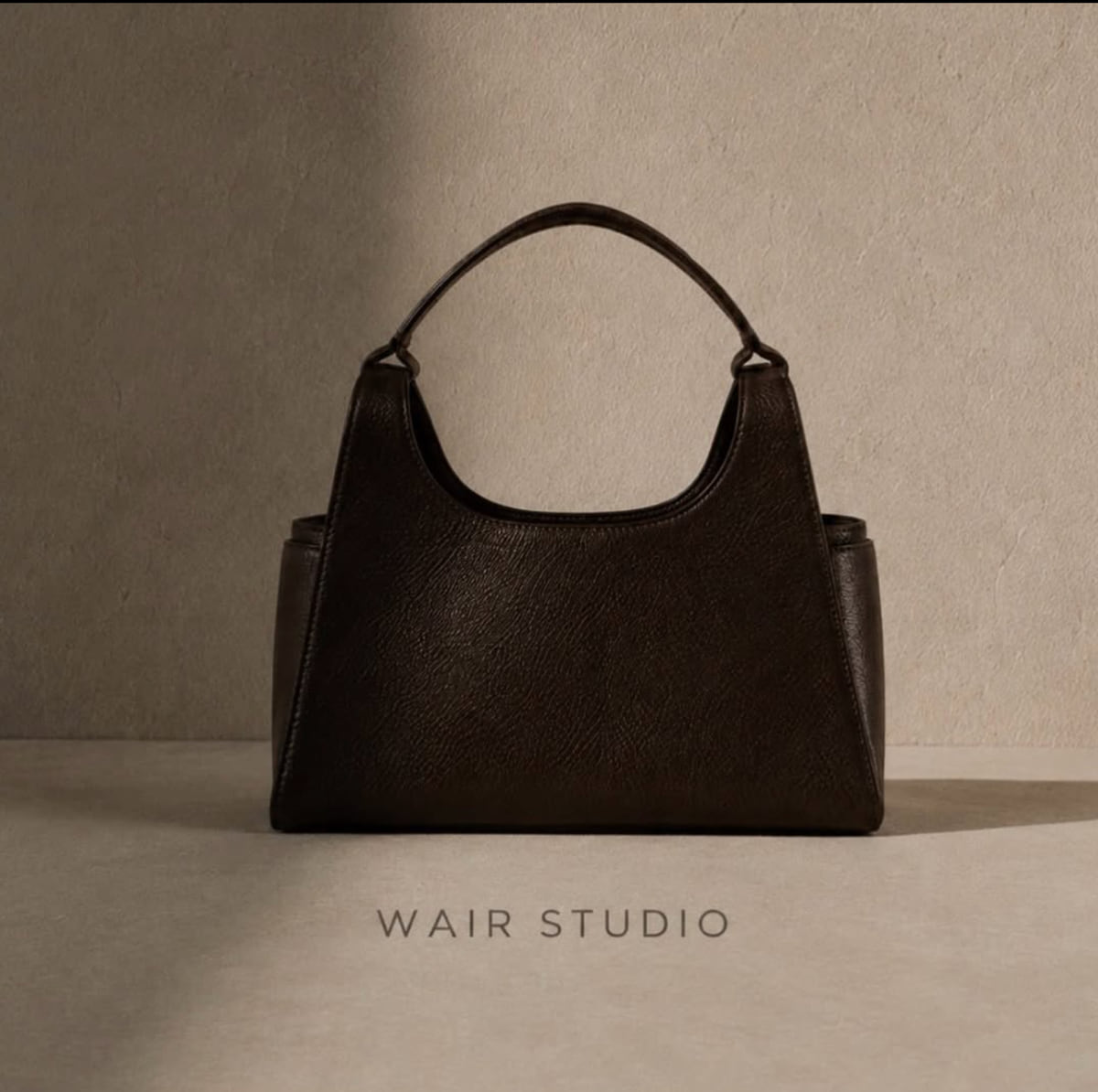 A dark brown pebbled leather top-handle bag photographed as a studio still life on a warm cream backdrop, the WAIR Studio wordmark below.
