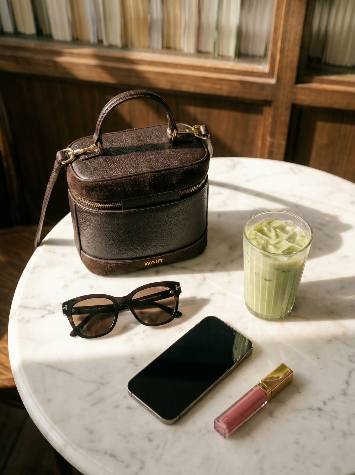 The Locket in espresso leather on a marble cafe table — sunglasses, an iced matcha, a phone, and a tinted lip gloss in the same frame