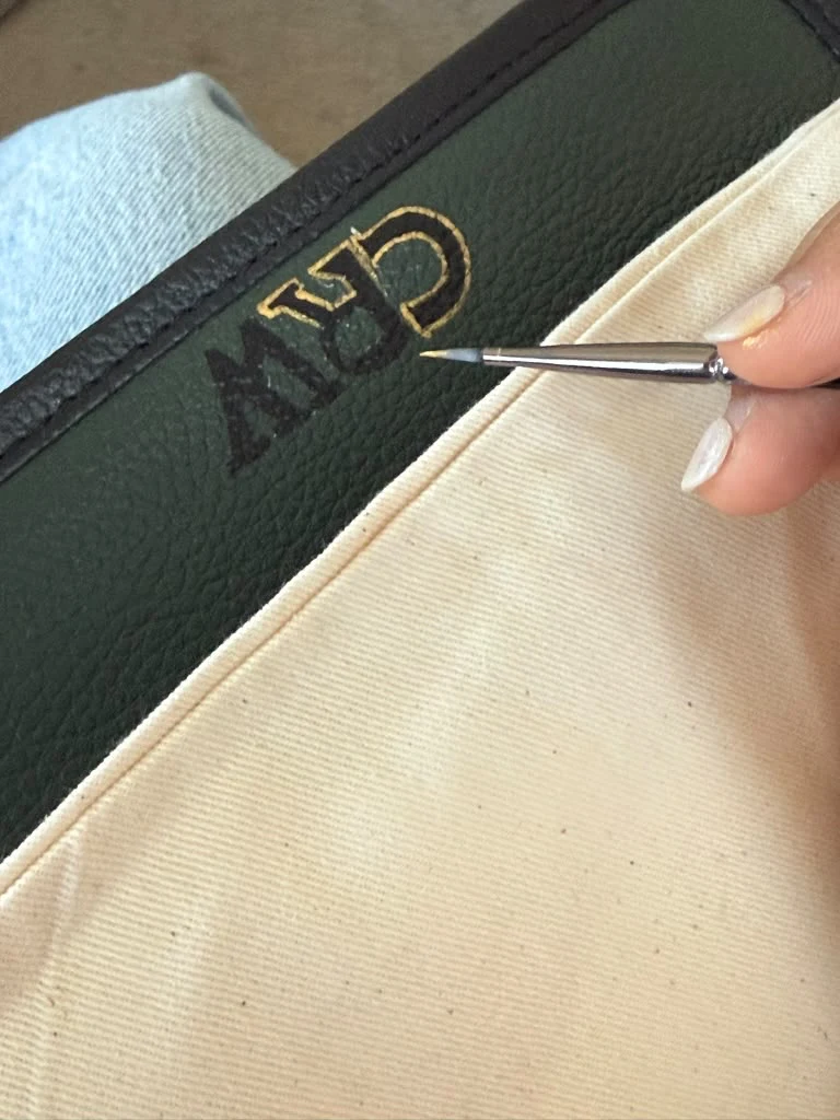 A fine brush adds a gold edge to dark initials on green leather.